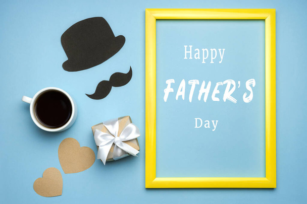 Σύνθεση ημέρας του πατέρα με κουτί δώρου, μαύρο μουστάκι, καπέλο, φλιτζάνι καφέ και καρδιά σε μπλε φόντο. Happy Father 's Day κείμενο σε κίτρινο πλαίσιο. Ευχετήρια κάρτα για τον μπαμπά. Πάνω όψη επίπεδη lay. - Φωτογραφία, εικόνα