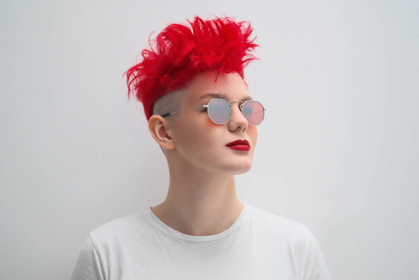 Portrait of a cool fashionable modern young girl. A short haircut with shaved temple. Dyed bright red hair. Red lipstick. Studio photo on white background. Suspenders and checkered pants. - Foto, Imagen