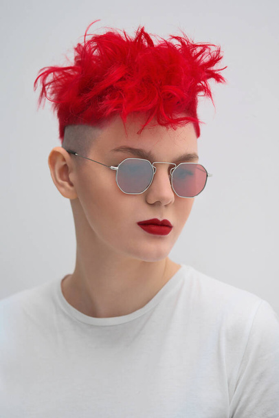 Portrait of a cool fashionable modern young girl. A short haircut with shaved temple. Dyed bright red hair. Red lipstick. Studio photo on white background. Suspenders and checkered pants. - Фото, изображение