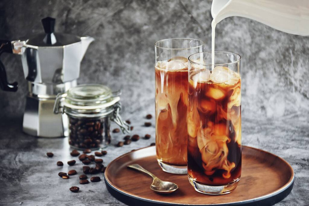 Ice coffee in a tall glass with cream poured over and coffee beans on a old rustic wooden board. Cold summer drink on a dark wooden background with copy space - 写真・画像