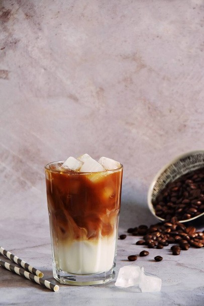 Ice coffee in a tall glass with cream poured over and coffee beans on a old rustic wooden board. Cold summer drink on a dark wooden background with copy space - Φωτογραφία, εικόνα