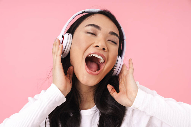 Cheerful pretty young asian woman listening to music with wireless headphones isolated over pink background, singing - Fotografie, Obrázek