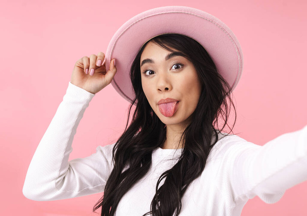 Cheerful pretty young asian woman taking a selfie while standing isolated over pink background, wearing hat - Fotografie, Obrázek