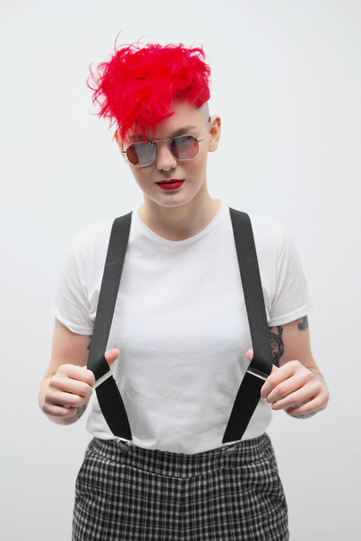 Portrait of a cool fashionable modern young girl. A short haircut with shaved temple. Dyed bright red hair. Red lipstick. Studio photo on white background. Suspenders and checkered pants. - Foto, Bild