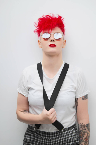 Portrait of a cool fashionable modern young girl. A short haircut with shaved temple. Dyed bright red hair. Red lipstick. Studio photo on white background. Suspenders and checkered pants. - Fotografie, Obrázek