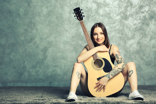 Vrouw met tatoeage en gitaar zittend op grijze achtergrond - Foto, afbeelding