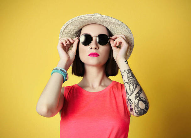 Belle jeune femme avec tatouage et lunettes de soleil portant un chapeau sur fond jaune
 - Photo, image