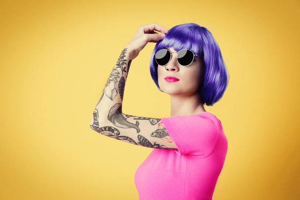 Gyönyörű fiatal nő tetoválás visel kék paróka és napszemüveg sárga háttér - Fotó, kép