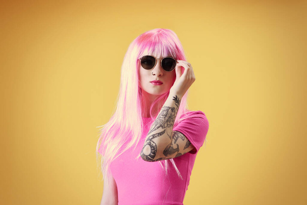 Mulher bonita com tatuagem vestindo peruca rosa e óculos de sol no fundo amarelo
 - Foto, Imagem