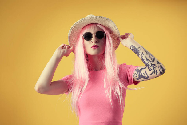 Hermosa mujer joven con tatuaje con peluca rosa, sombrero y gafas de sol sobre fondo amarillo
 - Foto, Imagen