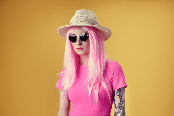 Beautiful young woman with tattoo wearing pink wig, hat and sunglasses on yellow background - Foto, Imagem