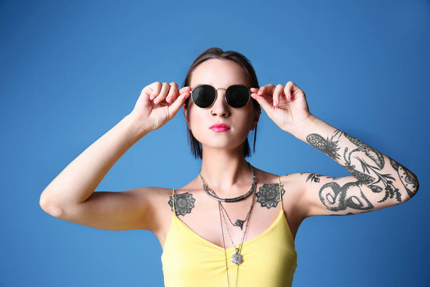 Beautiful young woman with tattoo wearing sunglasses and posing on blue background - Zdjęcie, obraz
