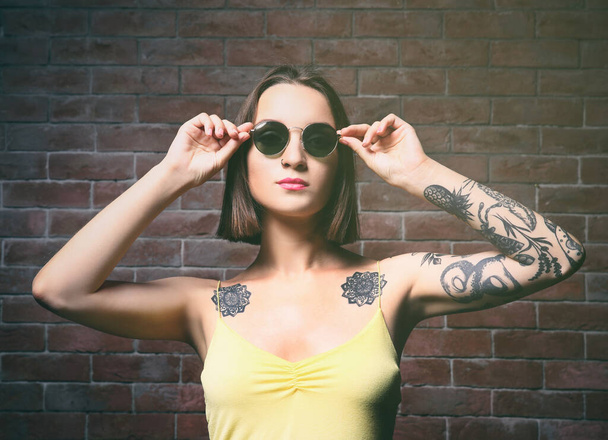 Mulher bonita com tatuagem usando óculos de sol e posando no fundo da parede de tijolo
 - Foto, Imagem
