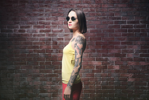 Kaunis nuori nainen, jolla on tatuointi aurinkolasit ja poseeraa tiiliseinän taustalla
 - Valokuva, kuva