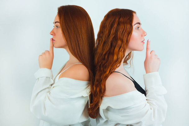 two redheaded young women both holding fingers near lips showing keep silence sign standing with their backs turned on isolated white backgroung, body language concept. - Fotó, kép