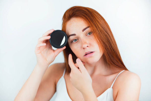 portrait of redheaded young woman looking natural holding face cream near her face standing on isolated white backgroung, care and beauty concept. - Fotó, kép