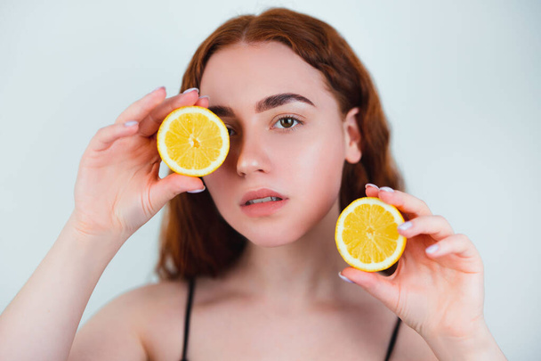 portrait of redheaded young woman standing on isolated white background holding half of orange in hand closing her eye with another half , beauty portrait concept. - Photo, image