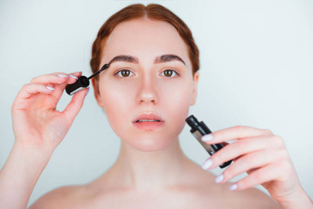 redheaded young woman looking natural applying brow mascara while doing makeup standing on isolated white background, makeup and beauty concept. - 写真・画像