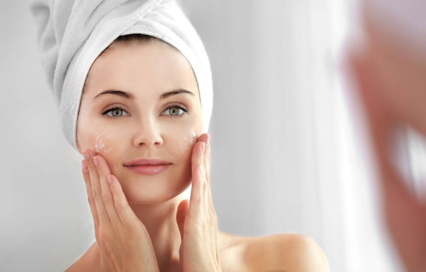 Mirror reflection of beautiful young woman with towel around head applying cream onto face - Фото, зображення