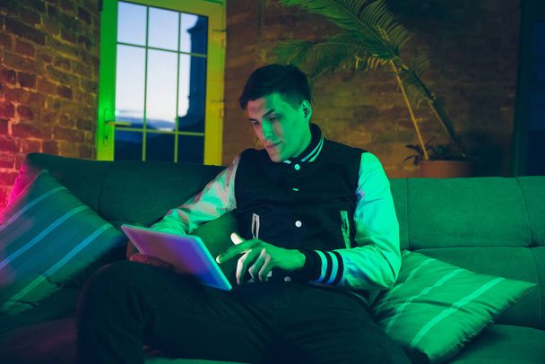 Cinematisch portret van knappe jongeman met behulp van apparaten, gadgets in neon verlichte interieur. Jeugdcultuur, felle kleuren - Foto, afbeelding