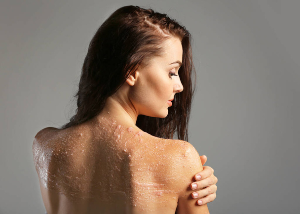 Young woman applying scrub on shoulder on grey background - 写真・画像