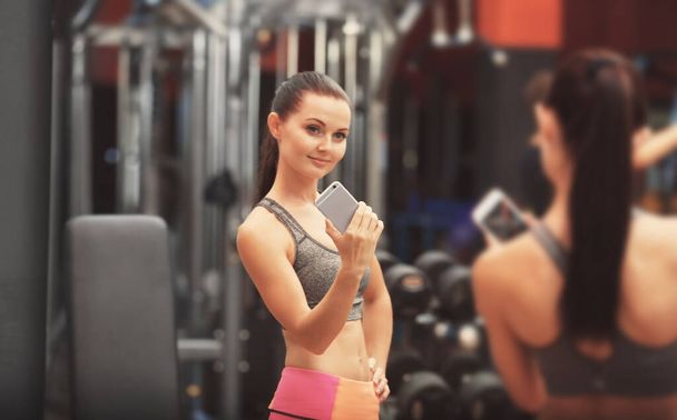 Νεαρή αθλητική γυναίκα που παίρνει selfie κοντά στον καθρέφτη στο γυμναστήριο - Φωτογραφία, εικόνα