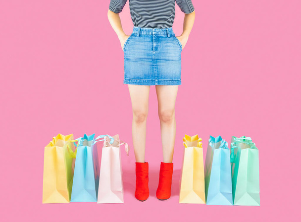 The legs of woman wore denim skirt and red boots standing with colorful pastel shopping bag on pink background  with copy space - Photo, image