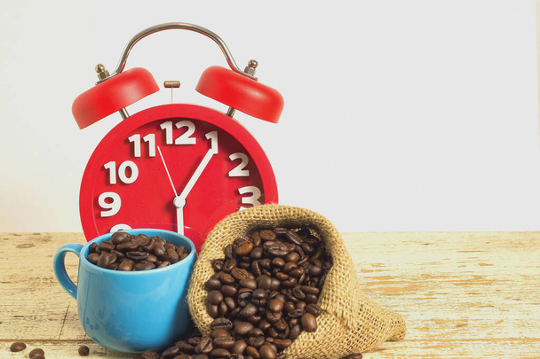 Relógio de alarme vermelho na mesa de madeira de fundo branco com grãos de café em um saco e xícara azul. Espaço de cópia para escrever texto
. - Foto, Imagem