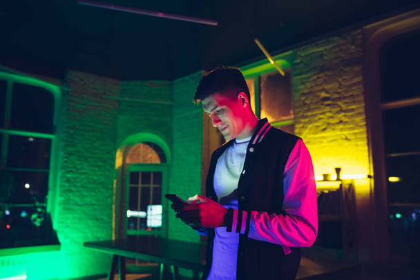 Cinematic portrait of handsome young man using devices, gadgets in neon lighted interior. Youth culture, bright colors - Fotografie, Obrázek