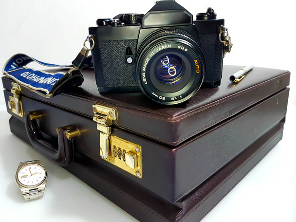 A black camera with lens, positioned on a brown briefcase with golden components, accompanied by a pen and a steel and gold colored watch. - Photo, Image