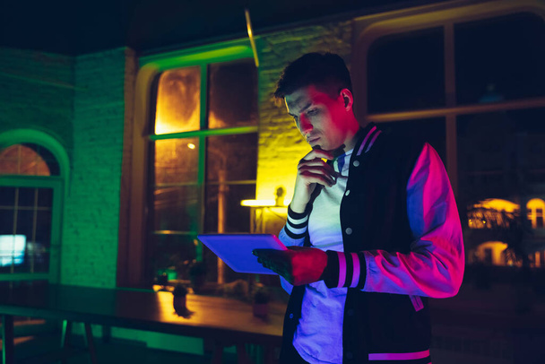 Cinematic portrait of handsome young man using devices, gadgets in neon lighted interior. Youth culture, bright colors - Фото, зображення