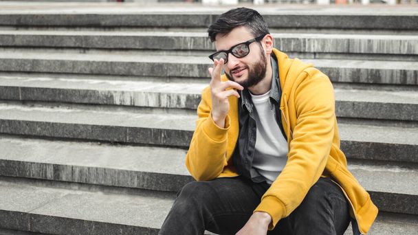 I choose you. Confident young man flirting sitting on steps outdoor.Handsome bearded millennial male in sunglasses and stylish clothes gesturing pointing fingers to camera,choice gesture, - Photo, Image