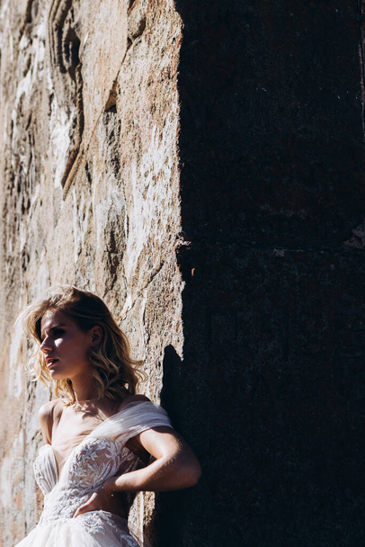 молодая невеста в длинном белом платье позирует для фото на фоне большой каменной стены, стильная фотография для продвижения женского салона одежды
 - Фото, изображение