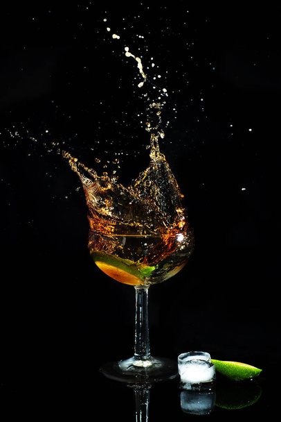Cocktail with cola and lime, which is splashed from a glass, on a black background. Drink on a black background - 写真・画像