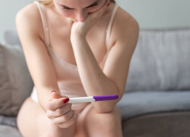 Single sad woman complaining holding a pregnancy test sitting on a couch in the living room at home - Foto, imagen