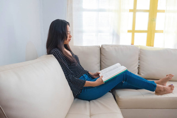 Μια γυναίκα κάθεται στον καναπέ διαβάζοντας ένα βιβλίο στο σαλόνι της. - Φωτογραφία, εικόνα