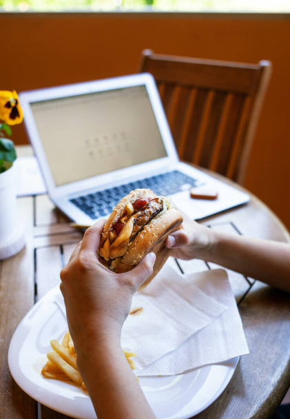 Close-up van de handen van een vrouw, tijdens het eten van een cheeseburger tijdens de lunchpauze, in de quarantaineperiode als gevolg van het coronavirus covid-19. Authentiek moment van slim werken met laptop in de thuisomgeving. - Foto, afbeelding