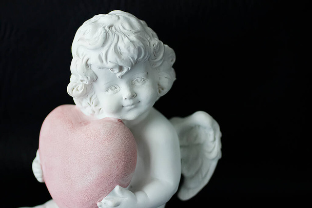 Anjo branco com um coração rosa nas mãos sobre um fundo preto. Foco suave
. - Foto, Imagem