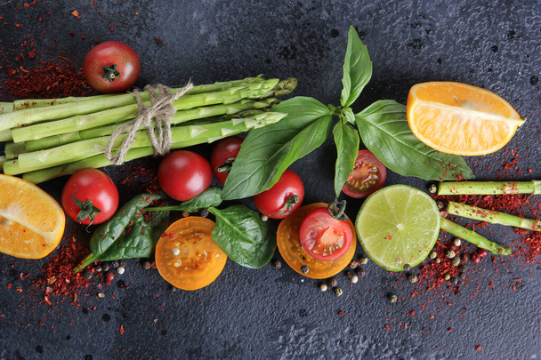 Fresh raw vegetables and herbs, asparagus, red cherry tomatoes, yellow tomatoes, lemon, lime, spinach, basil and spices on a black background. Top view, flatlay. Background image, copy space  - Foto, Bild
