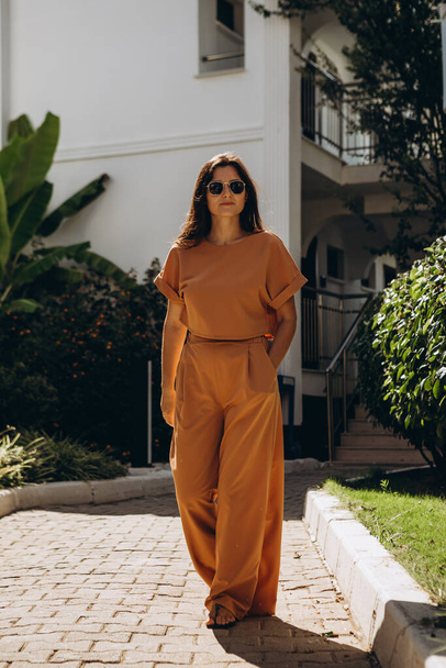 stylish young attractive woman wearing an orange-colored trouser suit and sunglasses photographed in the summer on a background of palm trees and the sea in the bright sun - Foto, Imagen