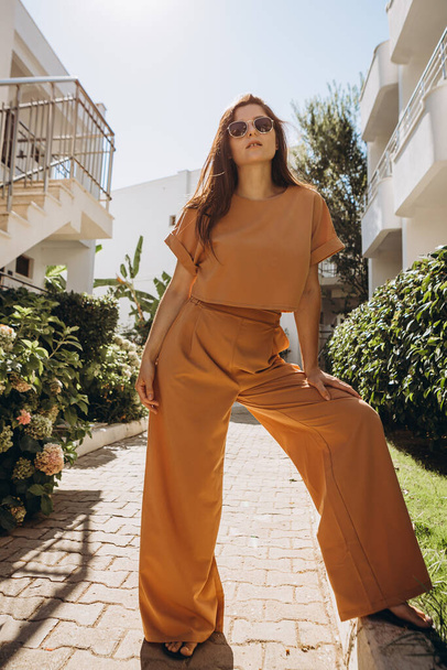 stylish young attractive woman wearing an orange-colored trouser suit and sunglasses photographed in the summer on a background of palm trees and the sea in the bright sun - Фото, изображение