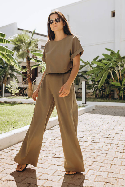 stylish young attractive woman dressed in dressed in a suit with a T-shirt and pants of olive color photographed in the summer on a background of palm trees and the sea in the bright sun - Zdjęcie, obraz