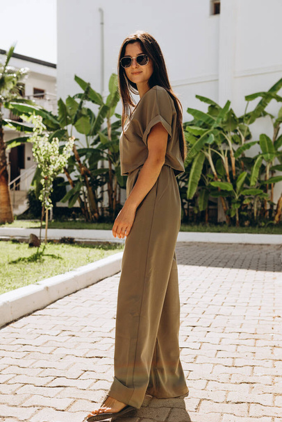 stylish young attractive woman dressed in dressed in a suit with a T-shirt and pants of olive color photographed in the summer on a background of palm trees and the sea in the bright sun - Zdjęcie, obraz