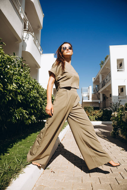 stylish young attractive woman dressed in dressed in a suit with a T-shirt and pants of olive color photographed in the summer on a background of palm trees and the sea in the bright sun - Φωτογραφία, εικόνα