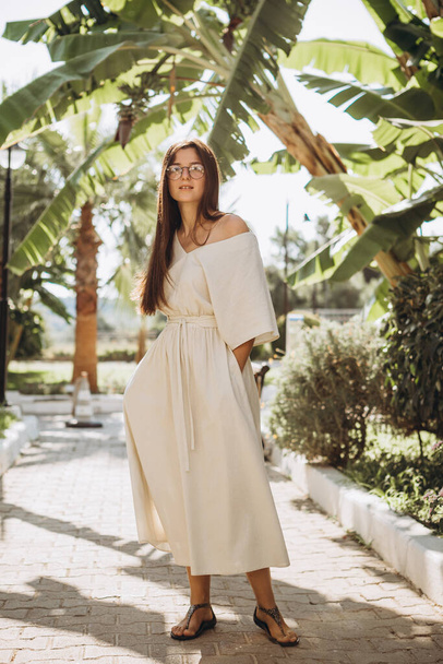 stylish young attractive woman dressed in a long linen dress with a hat photographed in the summer on a background of palm trees and the sea in the bright sun - 写真・画像