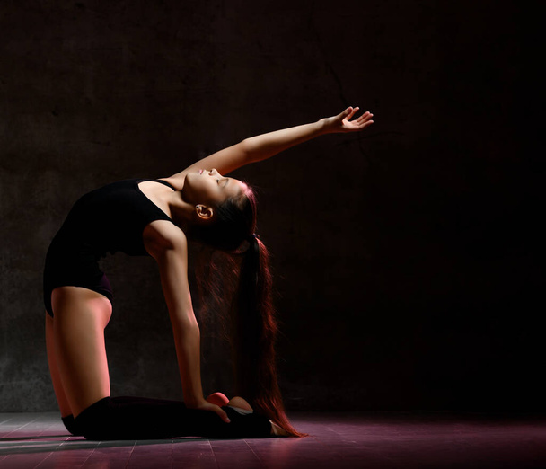 Young girl gymnast in black sport body and special footwear making gymnastic pose over dark background - Фото, зображення