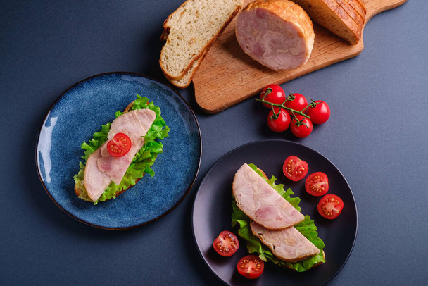 Sandwiches with turkey ham meat, green salad and fresh cherry tomatoes slices on black and blue plate near to ingredients on cutting board, blue minimal background, top view - Fotó, kép