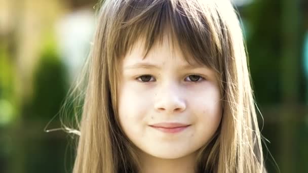 Porträt eines hübschen Mädchens mit grauen Augen und langen blonden Haaren, die im Freien auf verschwommenem, grünem Hintergrund lächeln. Nettes weibliches Kind an einem warmen Sommertag draußen. - Filmmaterial, Video