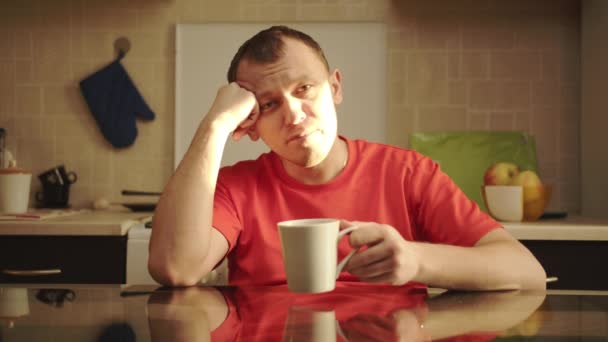 Portret mężczyzny siedzącego przy stole w kuchni w jaskrawo nasłonecznionej herbacie i patrzącego w kamerę - Materiał filmowy, wideo