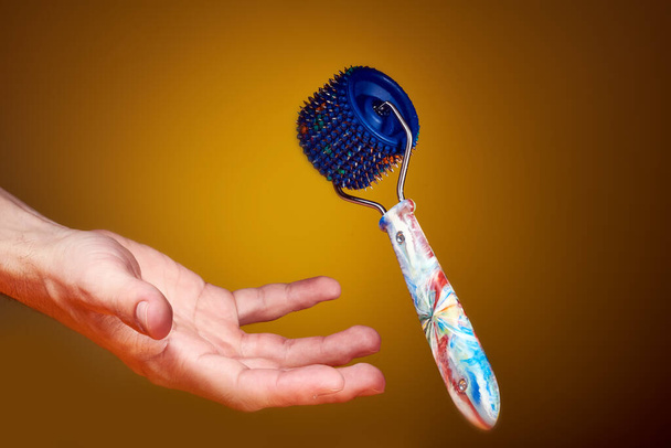 Massagegerät mit blauer Trommel, mehrfarbigem Kunststoffgriff und Metallnadeln auf gelbem Hintergrund mit der Hand. ist unbegründet. Motivfoto. Für Entspannung und Gesundheit. - Foto, Bild